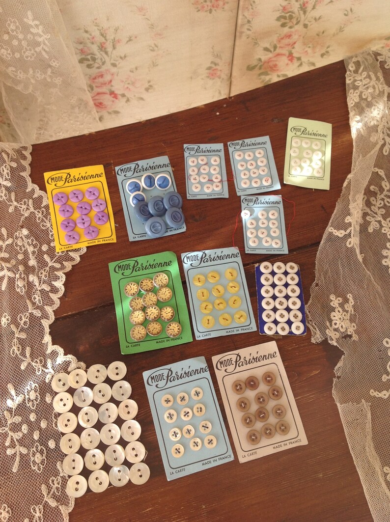 Op de afbeelding: Assortiment van vintage knoopkaarten met verschillende kleuren en patronen. De kaarten bevatten de tekst "Mode Parisienne" en "La Carte" met een blauwe, groene en gele achtergrond. De knoppen zijn gemaakt in Frankrijk.