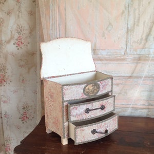 May include: A small, three-drawer chest made of cardboard covered in a floral patterned fabric. The chest has a curved top and is painted in a light beige color. The drawers have metal handles and are open, revealing the interior.