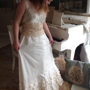 May include: A vintage-style white dress with a lace-trimmed bodice and a ruffled hem. The dress features a beige lace belt and spaghetti straps. The image is set in a room with a white piano and a floral rug.