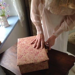 Puede incluir: Una caja rectangular con un estampado floral en tonos rosa y beige se encuentra sobre una mesa de madera oscura. La caja está siendo abierta por una persona que lleva un vestido de color claro. Un jarrón con flores es visible en el fondo.