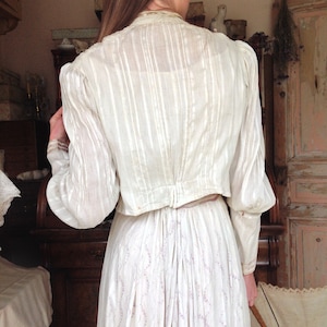 Puede incluir: Una blusa blanca de manga larga con cuello fruncido y espalda fruncida. La blusa est&aacute; hecha de una tela transparente con rayas verticales. La blusa se lleva sobre una falda blanca con estampado floral.
