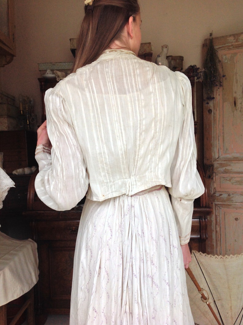 Puede incluir: Una blusa blanca de manga larga con cuello fruncido y espalda fruncida. La blusa est&aacute; hecha de una tela transparente con rayas verticales. La blusa se lleva sobre una falda blanca con estampado floral.