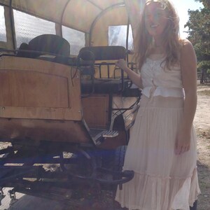 May include: A vintage covered wagon with a wooden body and canvas top. The wagon is brown and tan, with black metal accents. A person is standing next to the wagon wearing a cream-colored dress.