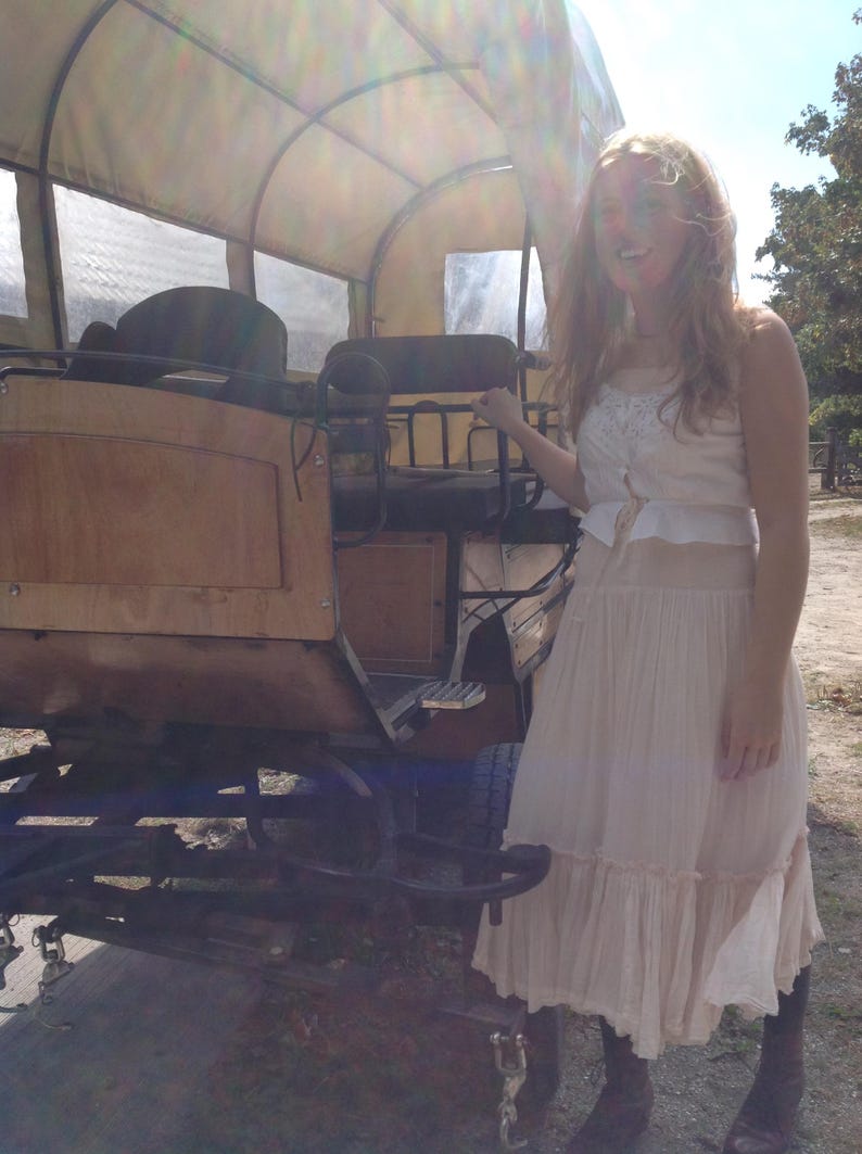 May include: A vintage covered wagon with a wooden body and canvas top. The wagon is brown and tan, with black metal accents. A person is standing next to the wagon wearing a cream-colored dress.