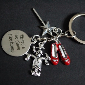 May include: Silver-toned keychain with charms featuring a star, a scarecrow, and a pair of red shoes. A round charm reads "There's no place like home." The keychain is on a dark background.