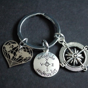 Può includere: Portachiavi argentato con tre ciondoli: una mappa del mondo a forma di cuore, un ciondolo rotondo con una bussola e la scritta "not all who wander are lost", e una rosa dei venti. Il portachiavi è su sfondo scuro.