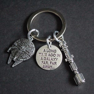 May include: Silver-toned keychain featuring three charms: a Millennium Falcon, a lightsaber, and a circular tag with the text "A LONG TIME AGO IN A GALAXY FAR, FAR AWAY...". The keychain is attached to a large metal ring.