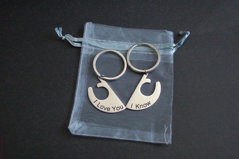 May include: Two silver keyrings, each with a unique shape that interlocks to form a whole. One keyring reads "I Love You," and the other reads "I Know." The keyrings are presented in a sheer, light grey drawstring bag.