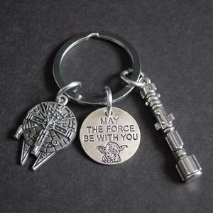 May include: A silver-coloured keyring with three charms: a Millennium Falcon, a round charm with Yoda and the text "MAY THE FORCE BE WITH YOU", and a lightsaber. The keyring is on a silver ring.