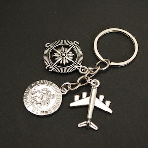 Op de afbeelding: Zilveren sleutelhanger met drie bedels: een kompas, een vliegtuig en een religieuze medaille met de woorden "Sint Christoffel Bescherm Ons".