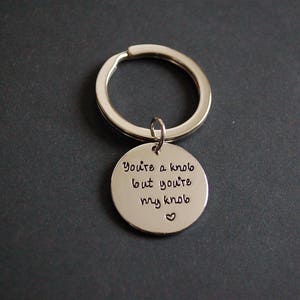 Op de afbeelding: Zilveren sleutelhanger met een ronde hanger met de tekst "You're a knob but you're my knob" met een klein hartje.