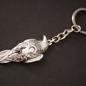 May include: Silver keyring featuring a detailed bird design with a crescent moon on its chest. The bird is lying on its back with its wings spread out.