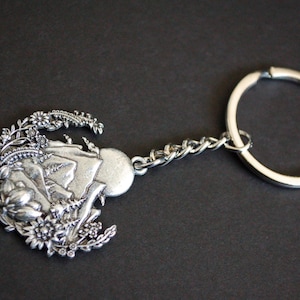 May include: A silver-toned metal keychain featuring a circular charm with a detailed embossed landscape design. The charm depicts a mountain range, a sun, and surrounding floral elements, connected to a standard circular split ring by a short chain.