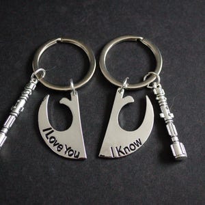 May include: Two silver keyrings with matching pendants. Each pendant is a crescent shape with the phrases "I Love You" and "I Know" engraved. Each keyring also has a small silver lightsaber charm.