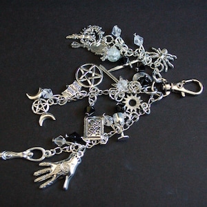 May include: A silver-toned metal keychain featuring multiple charms including a pentagram, a hand with an eye symbol, a tarot card, a spider, a dagger, crescent moons, a sun, and clear and black faceted beads attached to a lobster claw clasp.