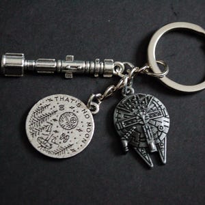 Peut inclure: Porte-clés argenté avec trois breloques : un sabre laser, un charme rond avec le texte "THAT'S NO MOON", et un charme du Faucon Millenium. Le porte-clés est sur un anneau argenté, idéal pour les clés ou les sacs.