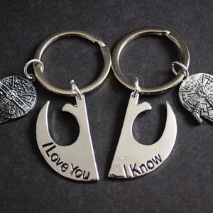 Puede incluir: Dos llaveros plateados con las palabras "I Love You" y "I Know" en ellos. Cada llavero tiene un dije plateado con forma de nave espacial.