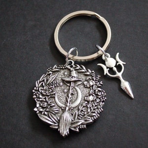 May include: Silver-toned keychain featuring a circular charm with a witch on a broomstick, a crescent moon, and floral details. A second charm with a triple moon symbol is attached to the keychain.