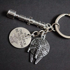 May include: Silver-toned keychain featuring a lightsaber, a Millennium Falcon charm, and a circular tag with the text "MAY THE FORCE BE WITH YOU" and a Yoda graphic. The keychain is on a dark background.