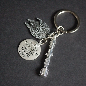 May include: Silver-coloured keychain with a Millennium Falcon charm, a lightsaber charm, and a circular charm with the text "A long time ago in a galaxy far, far away...". The keychain is on a dark background.