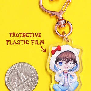 May include: A clear acrylic keychain featuring an anime character with brown hair, wearing a white hooded jacket with a red bow on top, blue jeans, and a white cat ear headband. The keychain has a rose gold metal clasp.