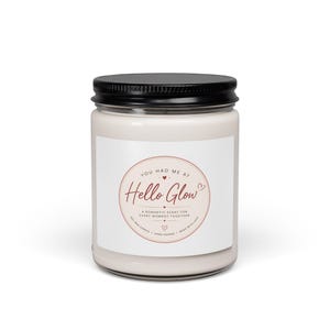 Puede incluir: Una vela blanca en un tarro de cristal transparente con tapa negra. La etiqueta presenta el texto "YOU HAD ME AT Hello Glow" en un diseño circular con un corazón. La vela se describe como una fragancia romántica para cada momento juntos.