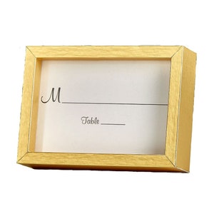 May include: Gold-colored table number frame with a white card insert. The card has the letter "M" and the word "Table" printed on it.