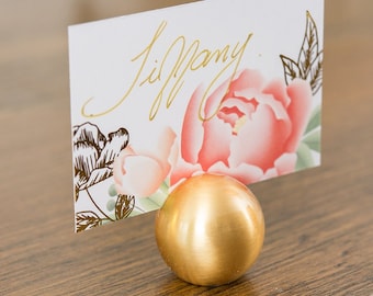 Puede incluir: Porta tarjetas de lugar de metal dorado con base redonda. Una tarjeta blanca con el nombre "Tiffany" escrito en letras doradas se muestra en el soporte. La tarjeta tiene un diseño floral con flores rosas y verdes.