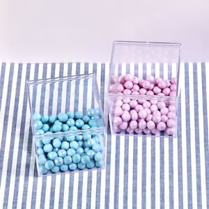 May include: Two clear plastic square boxes filled with candy. The box on the left contains blue candy and the box on the right contains pink candy. The boxes are sitting on a blue and white striped surface.