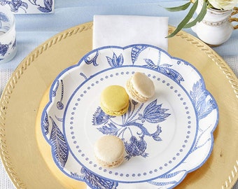 May include: A white paper plate with a blue and white floral design. The plate is on top of a gold charger plate. There are three macarons on the plate, two are beige and one is yellow.