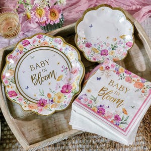 May include: A collection of floral-themed party supplies, including plates and napkins. The plates have a scalloped edge and gold trim, with the words "Baby in Bloom" in gold script. The napkins also feature the same text and floral design.