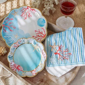 May include: A collection of coastal-themed paper plates and a matching napkin set. The plates feature a scalloped edge, gold trim, and illustrations of coral, starfish, and seashells. The napkin has blue and white stripes with a coral and starfish design.