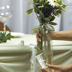 May include: White plastic table clip with a white plastic vase insert for holding a bouquet of flowers. The vase insert is being placed into the table clip.