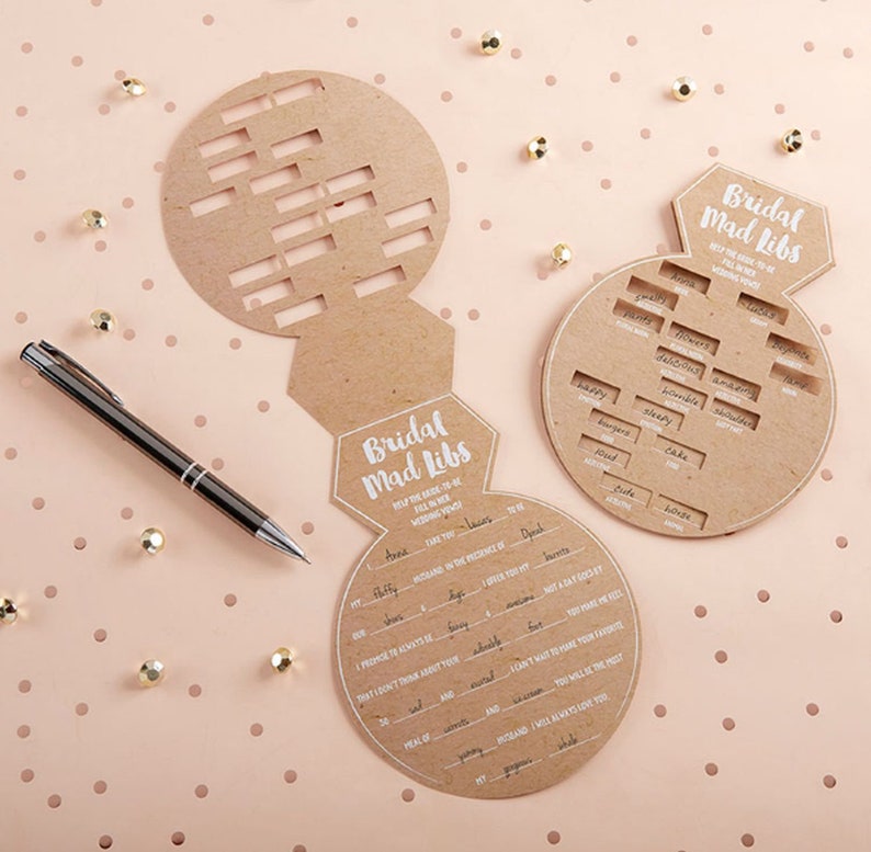 K&ouml;nnte beinhalten: Zwei braune Spielkarten aus Karton in Ringform mit dem Text "Bridal Mad Libs" und L&uuml;ckentexten &uuml;ber eine Hochzeit. Die Karten befinden sich auf einem rosa Hintergrund mit wei&szlig;en Punkten.