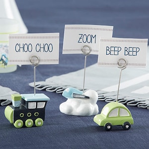 May include: Three table card holders with different transportation themes. A black and green train with the words "Choo Choo" on the card, a white cloud with a blue airplane and the word "Zoom" on the card, and a green car with the words "Beep Beep" on the card.