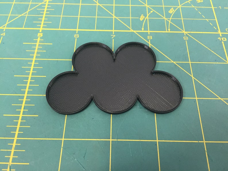 May include: A black plastic cloud-shaped object with five rounded sections. The object is on a green cutting mat with yellow lines.
