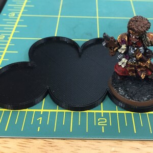 May include: A black plastic base with three connected circles, designed for tabletop gaming miniatures. The base is approximately 2 inches in diameter.