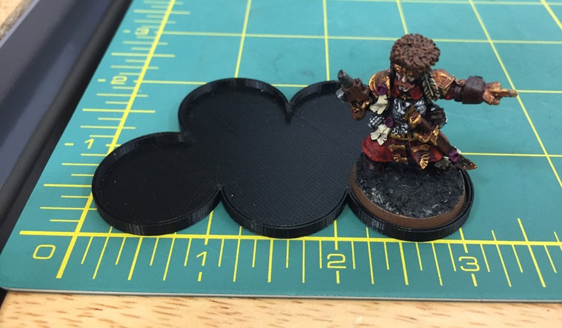 May include: A black plastic base with three connected circles, designed for tabletop gaming miniatures. The base is approximately 2 inches in diameter.