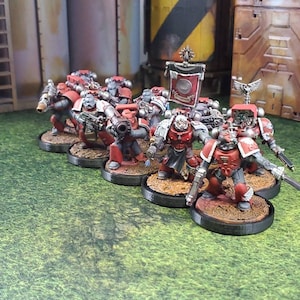 May include: A group of red and silver painted miniature figures, representing space marines, standing on circular bases. The figures are holding weapons and are posed in a battle formation.