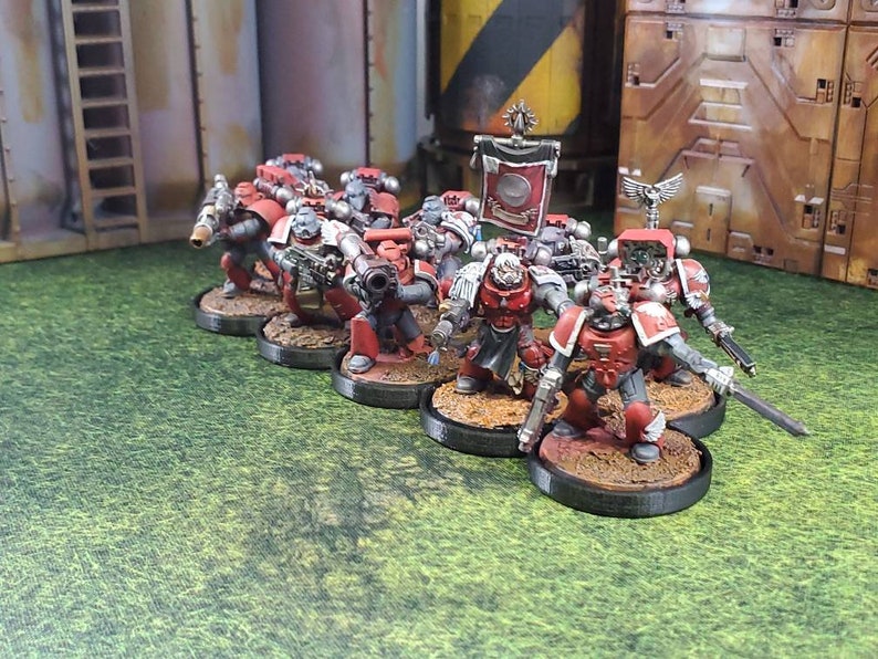 Op de afbeelding: Een groep rood en zilver geverfde miniatuurfiguren, die space marines voorstellen, die op ronde bases staan. De figuren houden wapens vast en staan in een gevechtsformatie.