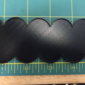 Op de afbeelding: Zwarte plastic basis met een gegolfde rand, ongeveer 12,7 cm lang. De basis is ontworpen om miniatuurfiguren of andere kleine voorwerpen te houden.