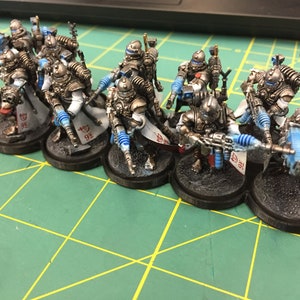 Op de afbeelding: Een groep van acht geverfde miniatuurfiguren in zilver en wit met blauwe accenten. De figuren houden wapens vast en staan op zwarte bases. De figuren bevinden zich op een groene ondergrond.