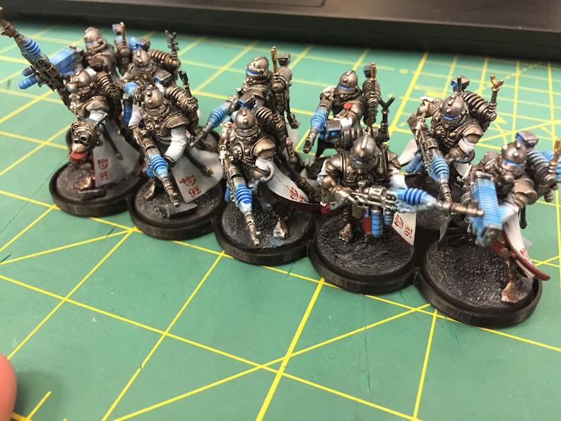 Op de afbeelding: Een groep van acht geverfde miniatuurfiguren in zilver en wit met blauwe accenten. De figuren houden wapens vast en staan op zwarte bases. De figuren bevinden zich op een groene ondergrond.