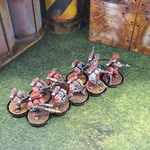 Op de afbeelding: Een groep rode en zilveren miniatuurfiguren met geweren, staande op zwarte bases. De figuren zijn in een lijn opgesteld, met het gezicht naar voren. De figuren zijn geplaatst tegen een groene gras achtergrond.