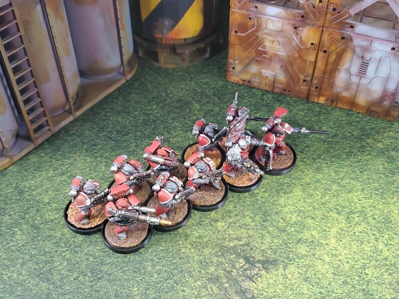 Op de afbeelding: Een groep rode en zilveren miniatuurfiguren met geweren, staande op zwarte bases. De figuren zijn in een lijn opgesteld, met het gezicht naar voren. De figuren zijn geplaatst tegen een groene gras achtergrond.