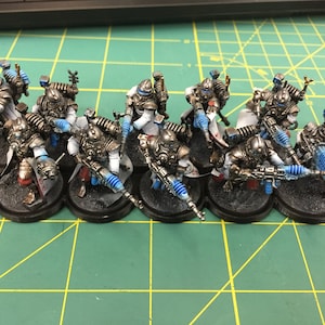 Op de afbeelding: Een groep van tien geverfde miniatuurfiguren in zilver, wit en blauw. De figuren houden wapens vast en staan op zwarte bases.