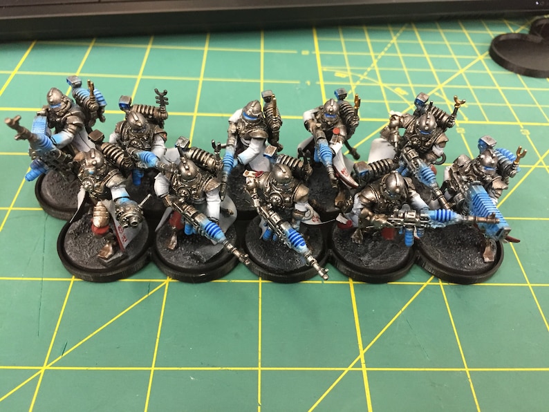Op de afbeelding: Een groep van tien geverfde miniatuurfiguren in zilver, wit en blauw. De figuren houden wapens vast en staan op zwarte bases.