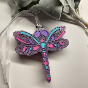 May include: A purple dragonfly ornament with pink, blue, and white accents. The dragonfly has a detailed design and is hanging from a white ribbon.