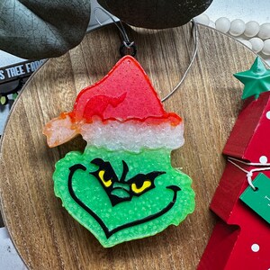 May include: Grinch Christmas ornament with a green face, yellow eyes, and a red Santa hat. The ornament is made of a glittery material and is hanging from a silver string.