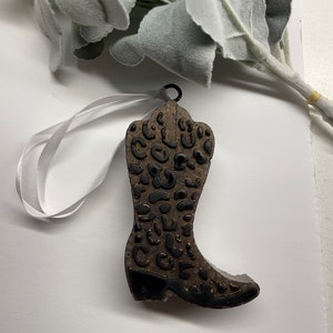 May include: A brown cowboy boot ornament with a black and gold leopard print design. The boot has a white ribbon attached to the top for hanging.